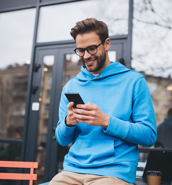 Middle aged man browses the internet on his cellphone while smiling outside on a bench with a coffee