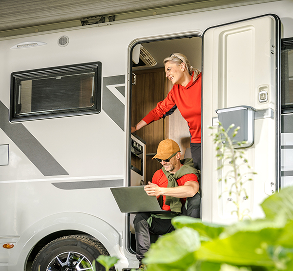 Grandmother talks through open RV door with a smile while relative works on laptop on RV steps