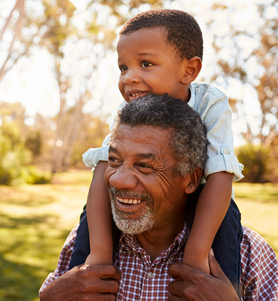 Retired man smiles as he carries grandchild on his shoulders for a piggyback ride outside