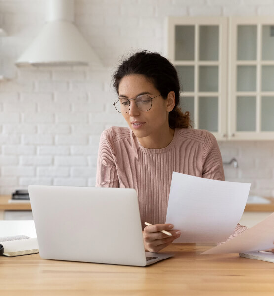 Lady with glasses looking at laptop and holding two sheets of paper.