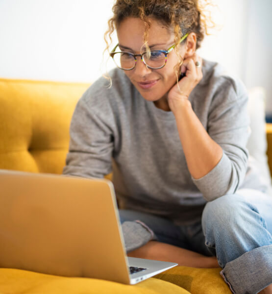 Lady with glasses on a yellow couch, browsing her laptop for savings options.