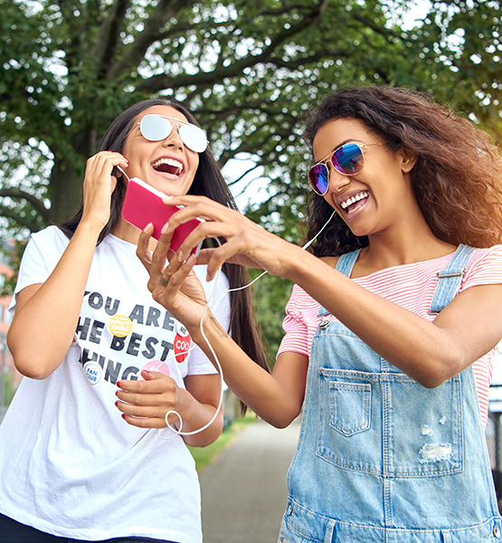 Young women laugh together while listen to music over a cellphone sharing earbuds with each other