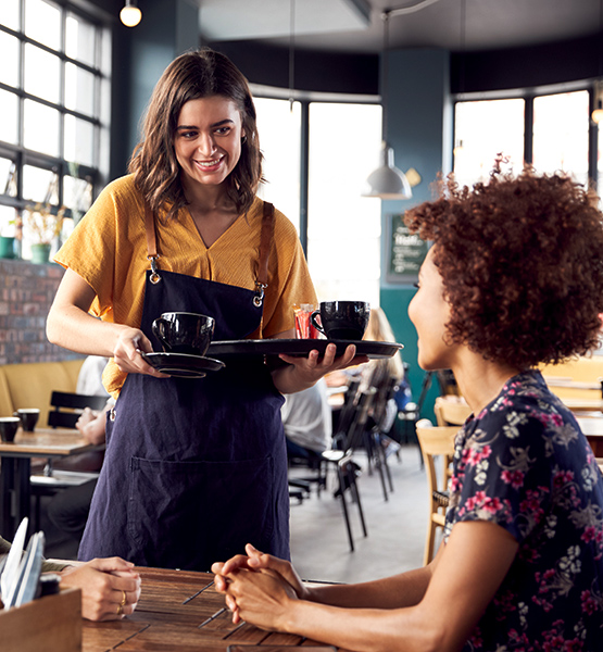 Young female waitress brings coffee order to waiting woman at a table in coffee shop