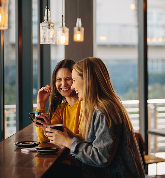 Two middle aged women chat and laugh together with mugs of coffee at a coffee shop