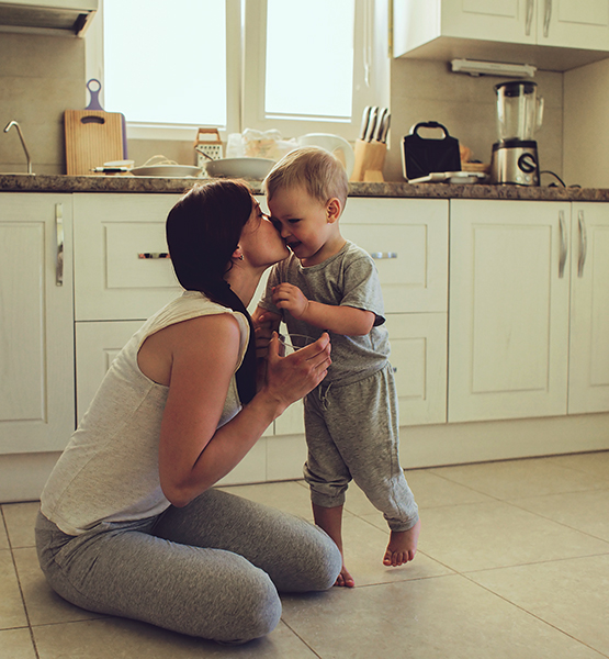 Young mother kisses smiling toddler son in kitchen of home