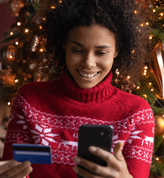 A young lady holding her phone and credit card sitting in front of a Christmas tree in a holiday sweater.