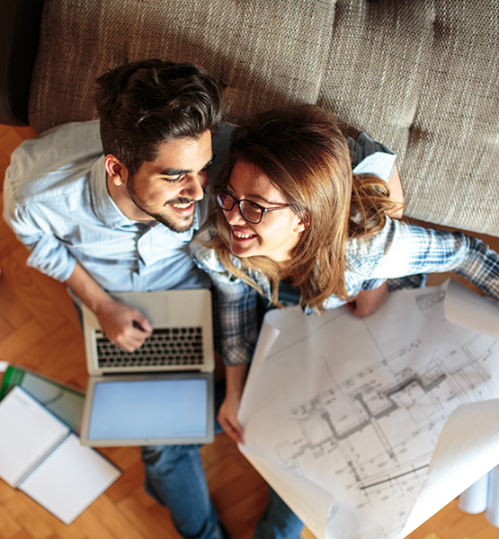Young man and woman giggling together on floor with laptop and house blueprints in front of couch
