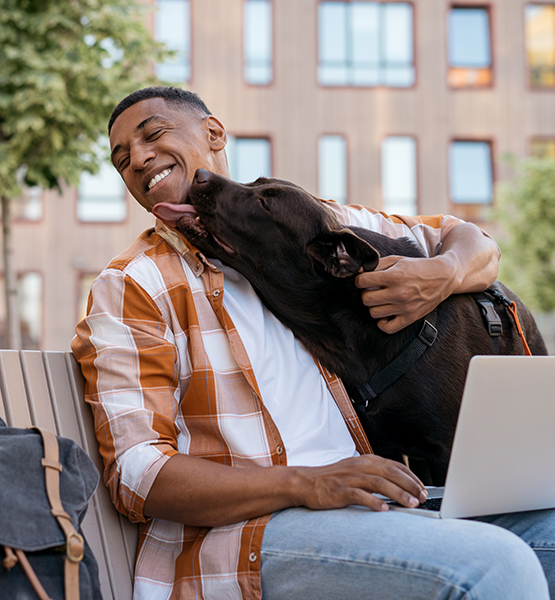 Man laughing and working on laptop while sitting on park bench outside while his dog licks his chin