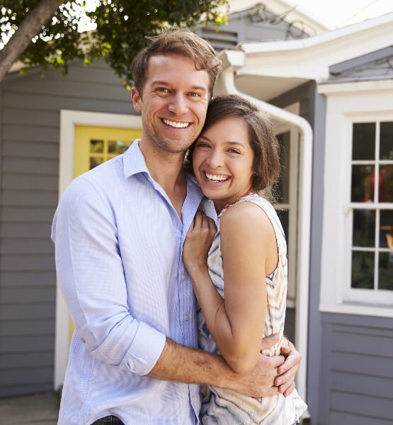 Happy Couple Smiling, next to a tree,  outside of a blue house. 