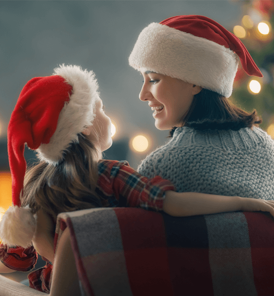 Mom and toddler in Santa hats sitting by Christmas tree.