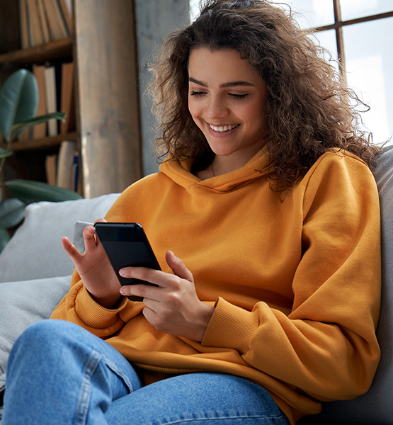 Middle aged woman sitting on couch at home while scrolling through her cellphone and smiling