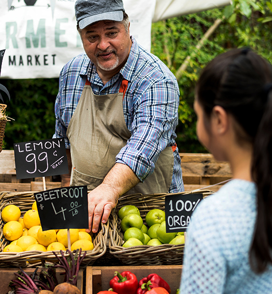 Business owner selling produce at farmers market with various fruit and vegetables talks to customer