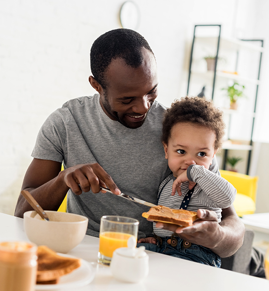 Father smiles with toddler son in lap while making a healthy breakfast at the kitchen counter