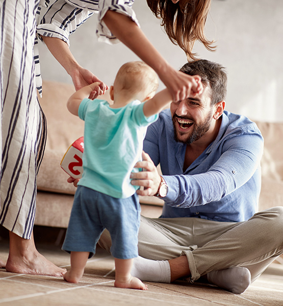 A father and mother laugh together helping infant son take his first steps in their living room home