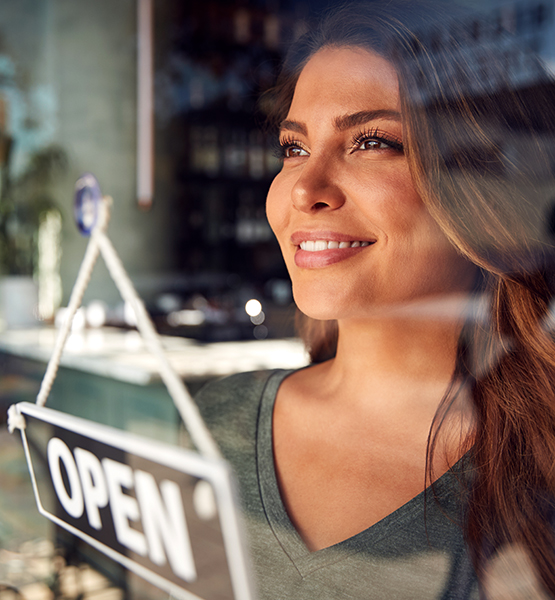 Female business owner looks out shop window smiling next to signage that says "open"