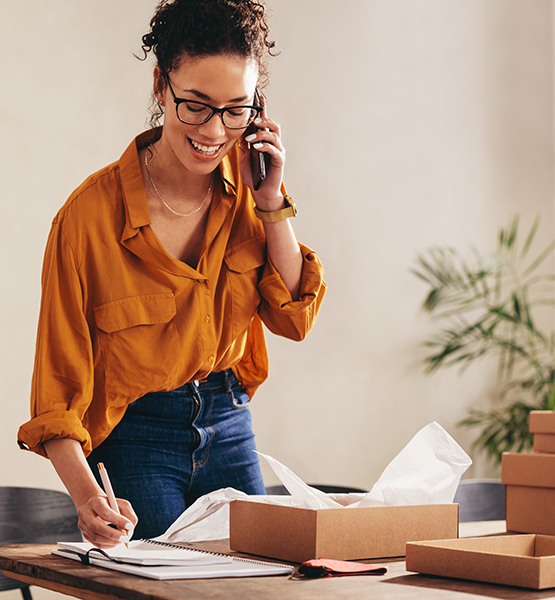 Business woman talks on phone with a smile while writing down order notes on a notepad at her desk